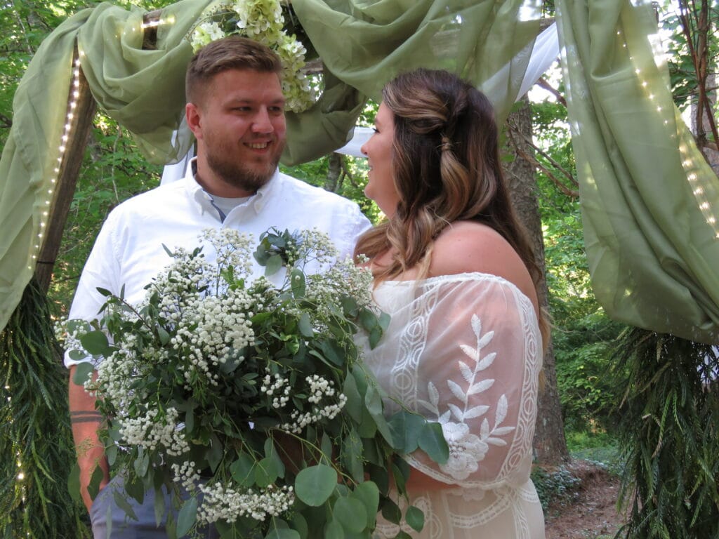 bride and groom looking at one another in front of a rustic wedding arch decorated in green drapes with lighting, bride is holding a bouquet of green mixed foliage trees and pines seen in the background