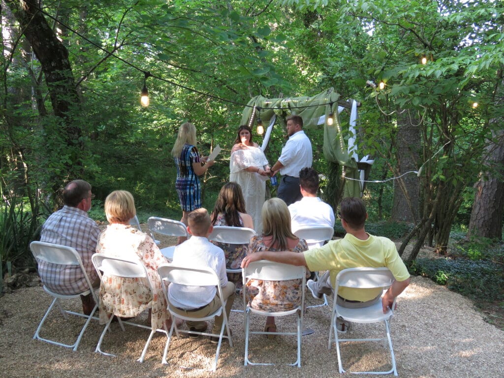 bride and groom facing minister during wedding while group of guest look on in front of a rustic wedding arch decorated in green drapes with lighting, bride is holding a bouquet of green mixed foliage trees and pines seen in the background