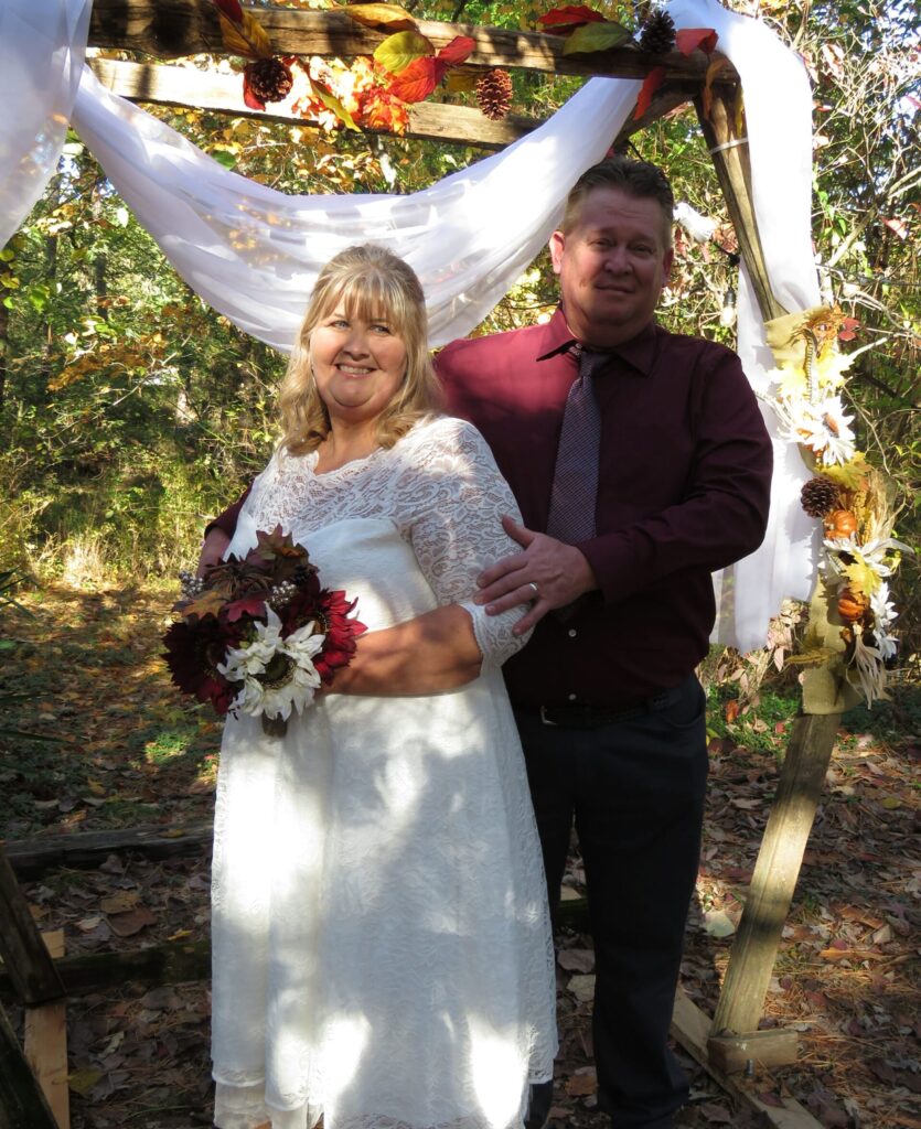 bride and groom in afternoon sun of fall standing under rustic wedding arch decorated in fall flower colors and white drapes