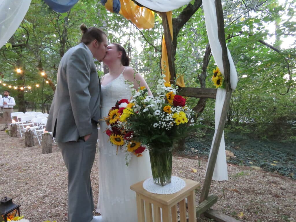 bride and groom kissing under rustic wedding arch with trees in the background