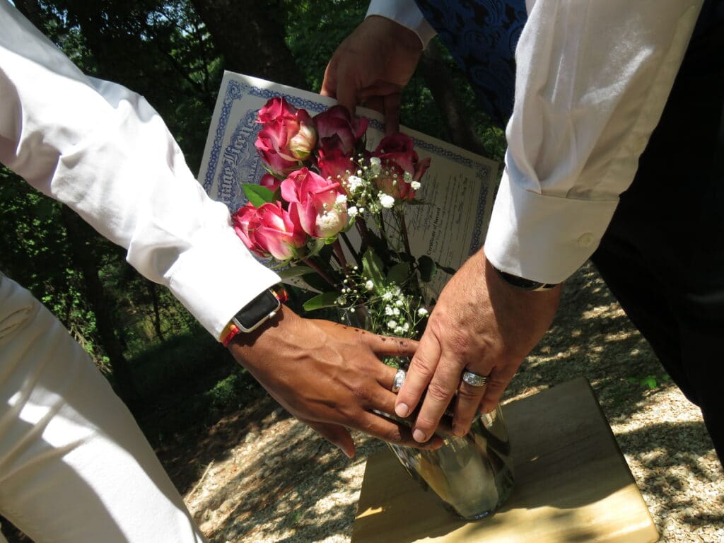 groom and groom hands with wedding bands together in front of flower bouquet with marriage license in background