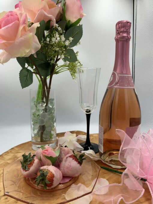 plate of pink chocolate covered strawberries next to bottle of pink champagne and vase of three pink roses