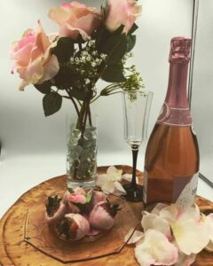 Plate with six pink chocolate covered strawberries next to bottle of pink Moscato champagne and vase of 3 pink roses 