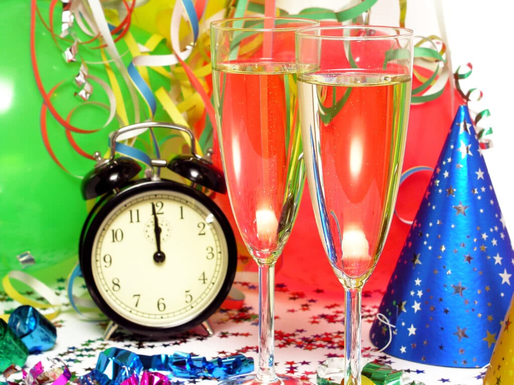 Two Champagne glasses next to a party hat and balloons and alarm clock pointed to midnight 