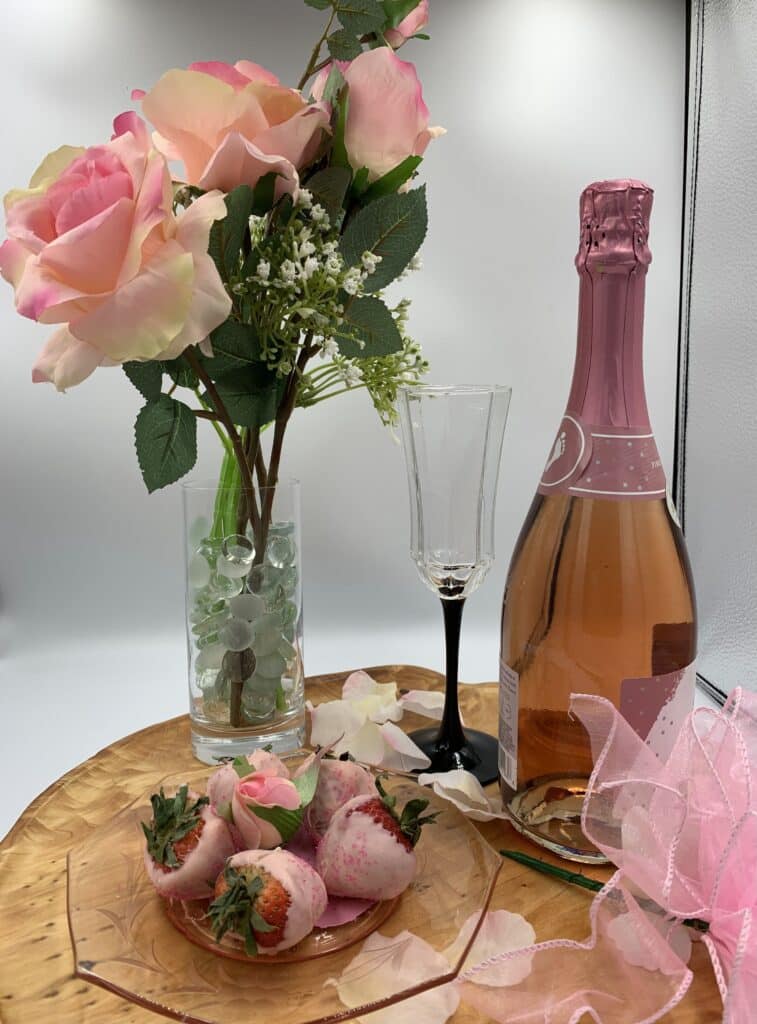 Three pink roses, bottle of pink champagne with a glass next to it, plate with strawberries dipped in pink chocolate, all sitting on a hard wood plate