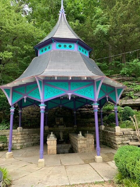 Purple and turquoise Gazebo sitting over a white brick fresh water spring surrounded by green forest.
