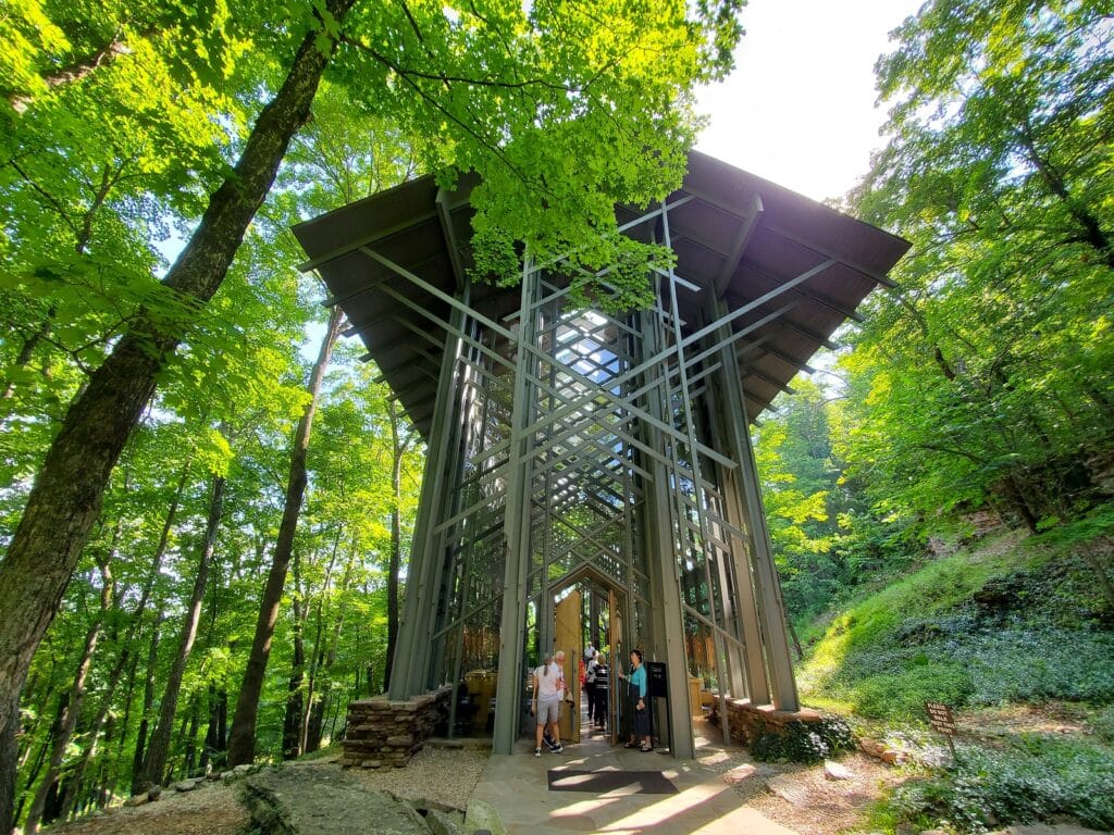 photo of glass chapel from outside, tall columns of glass with door and people standing in front of the door ready for a photo op. Area surrounded by a green forest