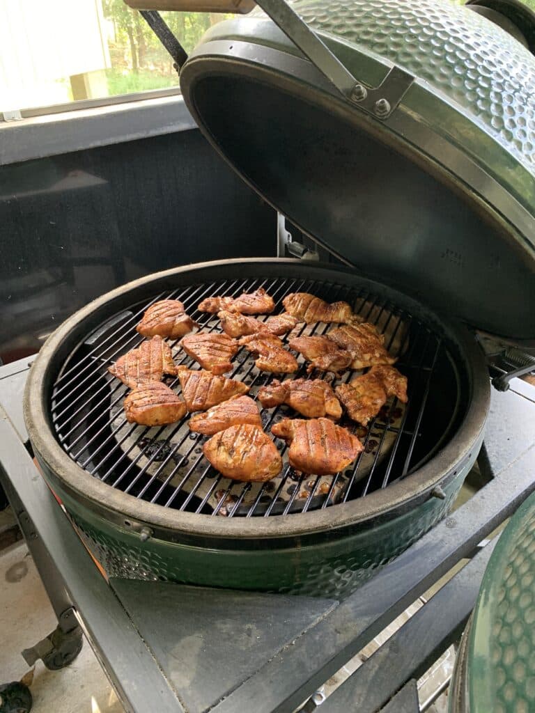 Several chicken thighs cooking on a grill with with the lid up and smoke curling around