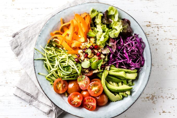 Vegan buddha bowl. Bowl with fresh raw vegetables - cabbage, carrot, zucchini, lettuce, watercress salad, tomatoes cherry and avocado, nuts and pomegranate. White background, top view